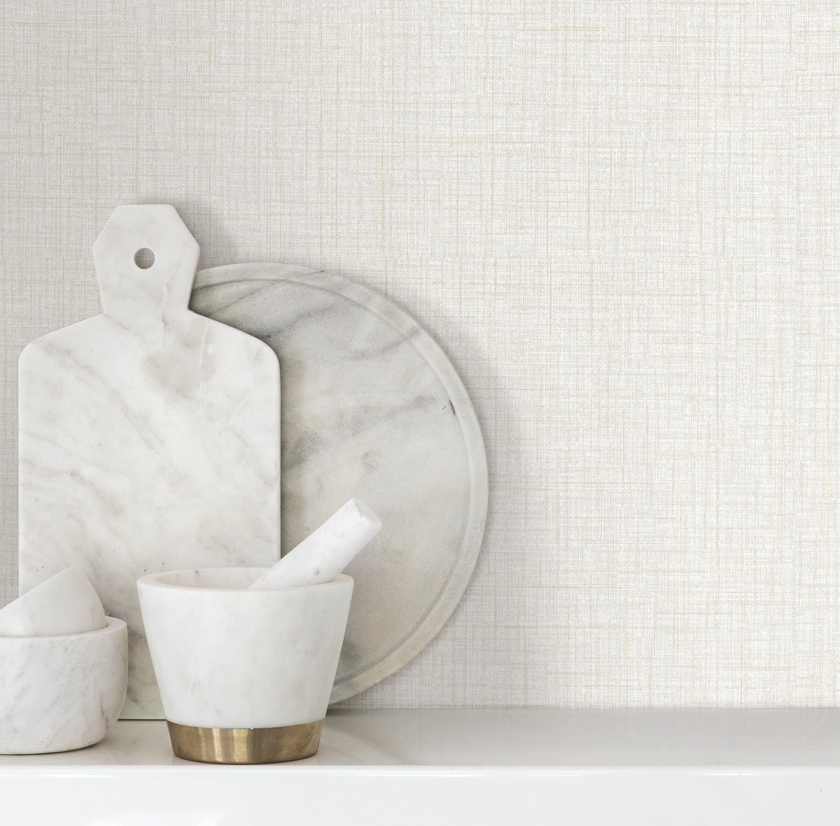 Beau Faux Fabric Wallpaper by York Wallcoverings serves as a light, textured backdrop for marble kitchenware, including a cutting board, round platter, and mortar and pestle arranged on a white surface.