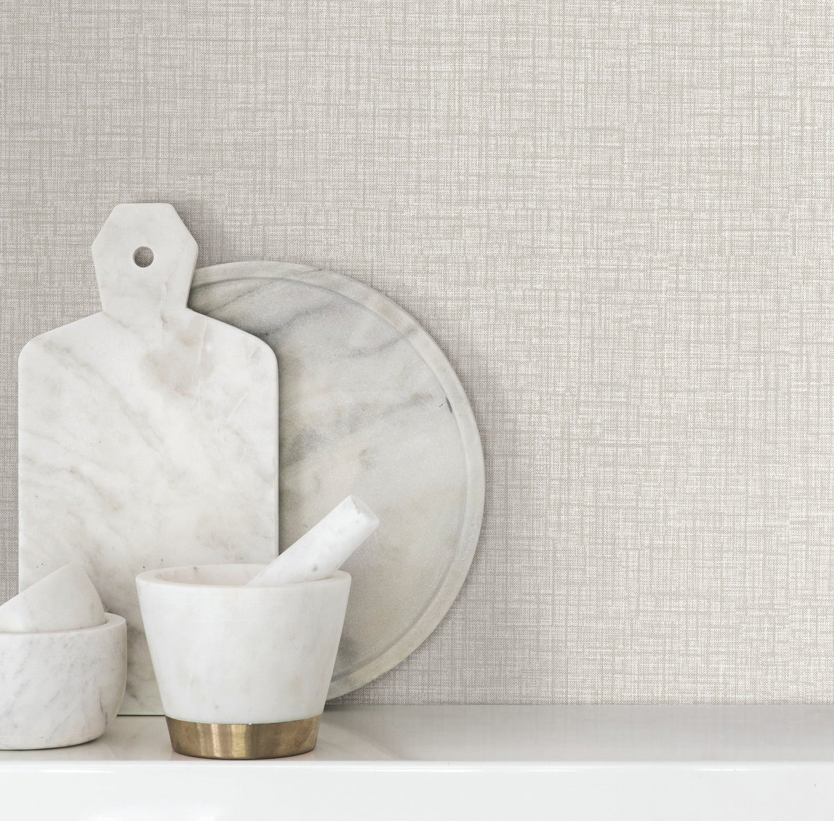 Beau Faux Fabric Wallpaper by York Wallcoverings serves as a textured beige backdrop behind a marble cutting board, round tray, and mortar and pestle arranged on a white shelf.