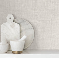 Beau Faux Fabric Wallpaper by York Wallcoverings serves as a textured beige backdrop behind a marble cutting board, round tray, and mortar and pestle arranged on a white shelf.