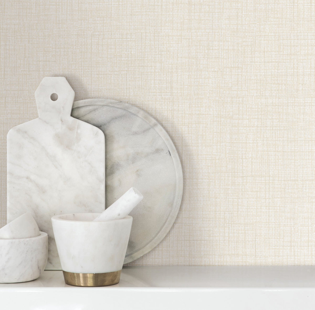 Beau Faux Fabric Wallpaper by York Wallcoverings serves as a backdrop for white marble kitchen items, including a cutting board, round tray, and mortar and pestle, arranged on a white countertop.