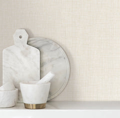 Beau Faux Fabric Wallpaper by York Wallcoverings serves as a backdrop for white marble kitchen items, including a cutting board, round tray, and mortar and pestle, arranged on a white countertop.