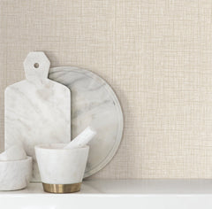 White marble kitchen items, including a cutting board, round tray, and mortar and pestle set, are arranged on a white surface against a beige Beau Faux Fabric Wallpaper backdrop by York Wallcoverings.