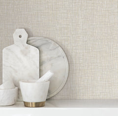 Beau Faux Fabric Wallpaper by York Wallcoverings serves as the backdrop for marble kitchen accessories, including a mortar and pestle, a round marble tray, and a marble cutting board with a handle, all arranged on a white surface.