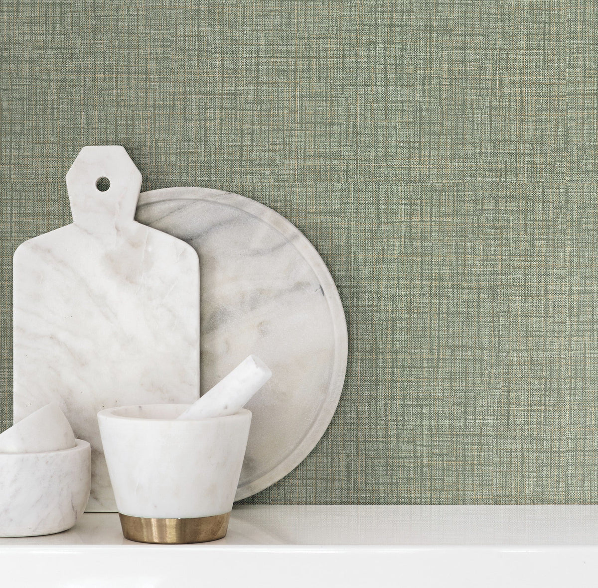 Beau Faux Fabric Wallpaper by York Wallcoverings creates a green textured backdrop behind marble cutting boards and a mortar and pestle arranged on a white surface.