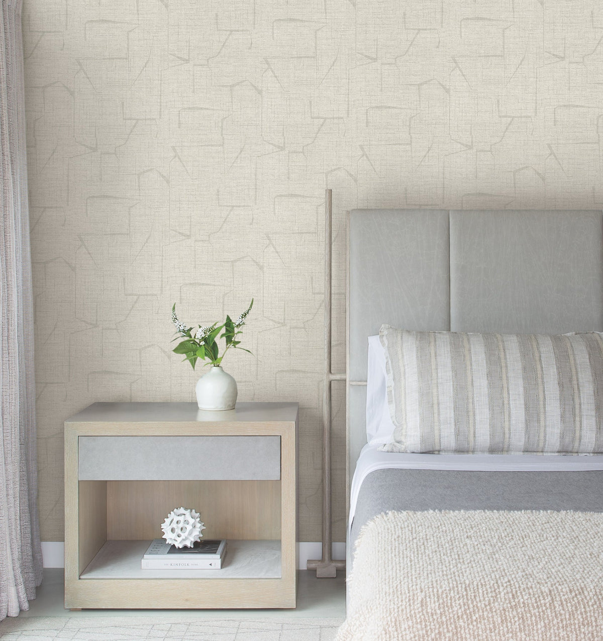 A modern bedroom featuring a light gray headboard, striped pillow, beige blanket, wooden nightstand with books, a white decorative object, and a vase with greenery is set against Lyle Abstract Geometric Wallpaper by York Wallcoverings.