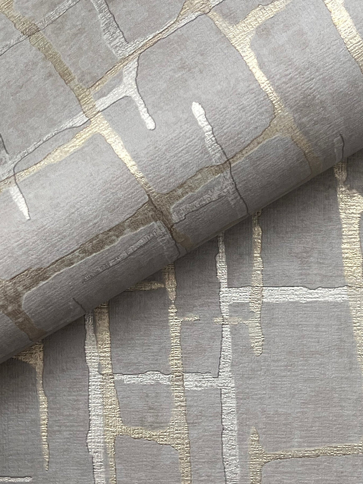 A close-up of the Rian Linear Abstract Wallpaper by York Wallcoverings showcases metallic gold and silver abstract grid patterns on textured grey paper, with one sheet partially rolled over another to highlight the wallpaper’s shimmering design.