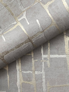 A close-up of the Rian Linear Abstract Wallpaper by York Wallcoverings showcases metallic gold and silver abstract grid patterns on textured grey paper, with one sheet partially rolled over another to highlight the wallpaper’s shimmering design.