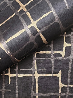 A close-up of two sheets of York Wallcoverings’ Rian Linear Abstract Wallpaper, featuring a black background with an abstract gold grid pattern and textured finish, with one sheet partially rolled over the other.