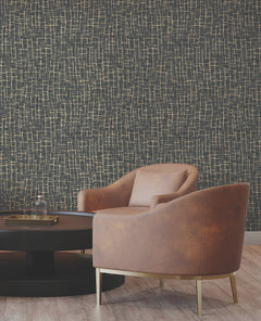 A modern brown leather chair with gold legs sits beside a round black coffee table. The background features York Wallcoverings’ Rian Linear Abstract Wallpaper in a dark hue with an abstract gold line pattern. The floor is light wood.