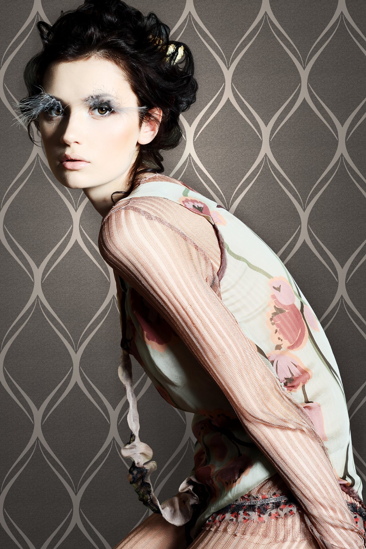 A woman in a floral top poses sideways against a wall adorned with York Wallcoverings’ Gustav Geometric Wallpaper, showcasing dramatic white feathered eyelashes and styled dark hair for a striking, avant-garde high-fashion look.