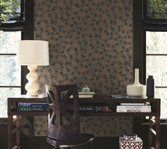 A wooden desk with books, a sculptural lamp, and vases sits against a wall adorned with York Wallcoverings’ Ophidia Wallpaper, featuring a geometric, chain-like pattern. A wooden chair is positioned in front of the desk as light filters through windows on both sides.