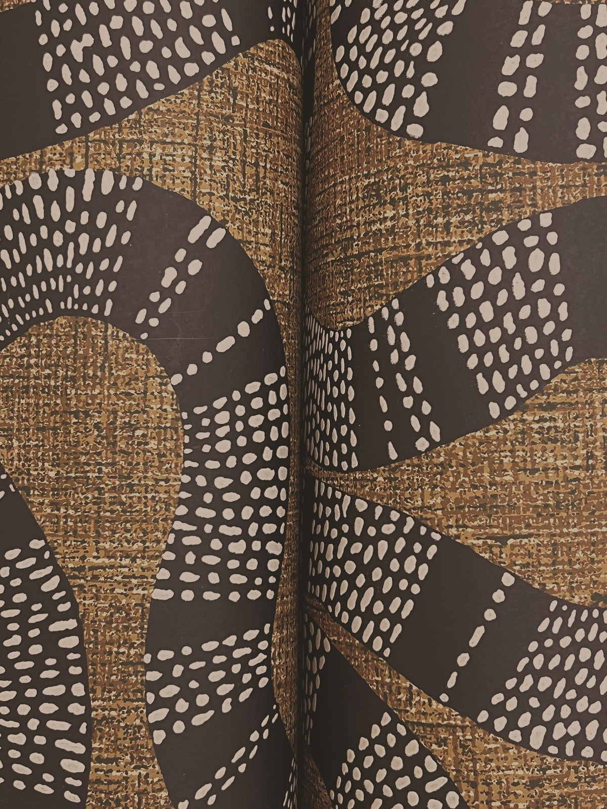 Ophidia Wallpaper by York Wallcoverings features curved black and white dotted lines forming abstract, snake-like patterns on a textured brown and gold background, creating a striking design that aligns seamlessly at the center.