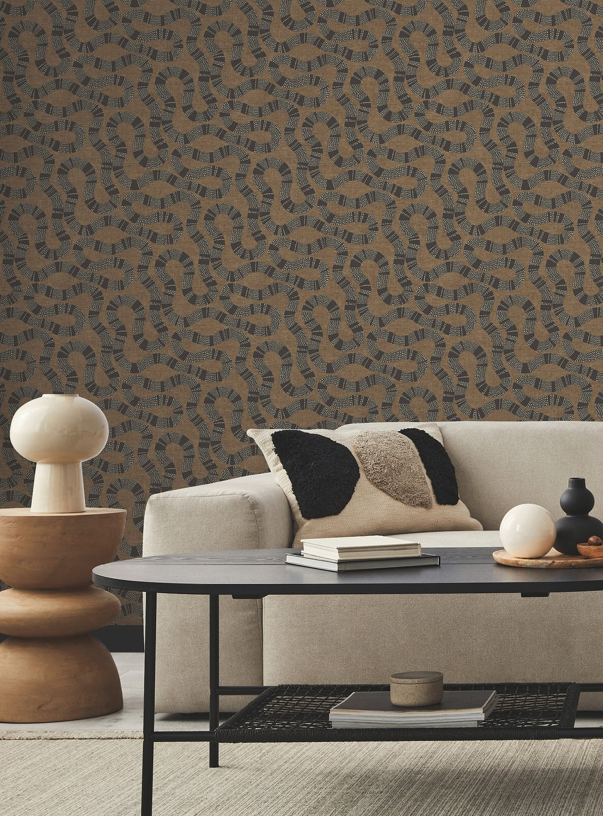 Modern living room with a beige sofa, monochrome textured pillows, round side table with cream lamp, black coffee table, and a beige carpet. The wall features Ophidia Wallpaper by York Wallcoverings in a brown and black abstract pattern.