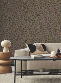 Modern living room with a beige sofa, monochrome textured pillows, round side table with cream lamp, black coffee table, and a beige carpet. The wall features Ophidia Wallpaper by York Wallcoverings in a brown and black abstract pattern.