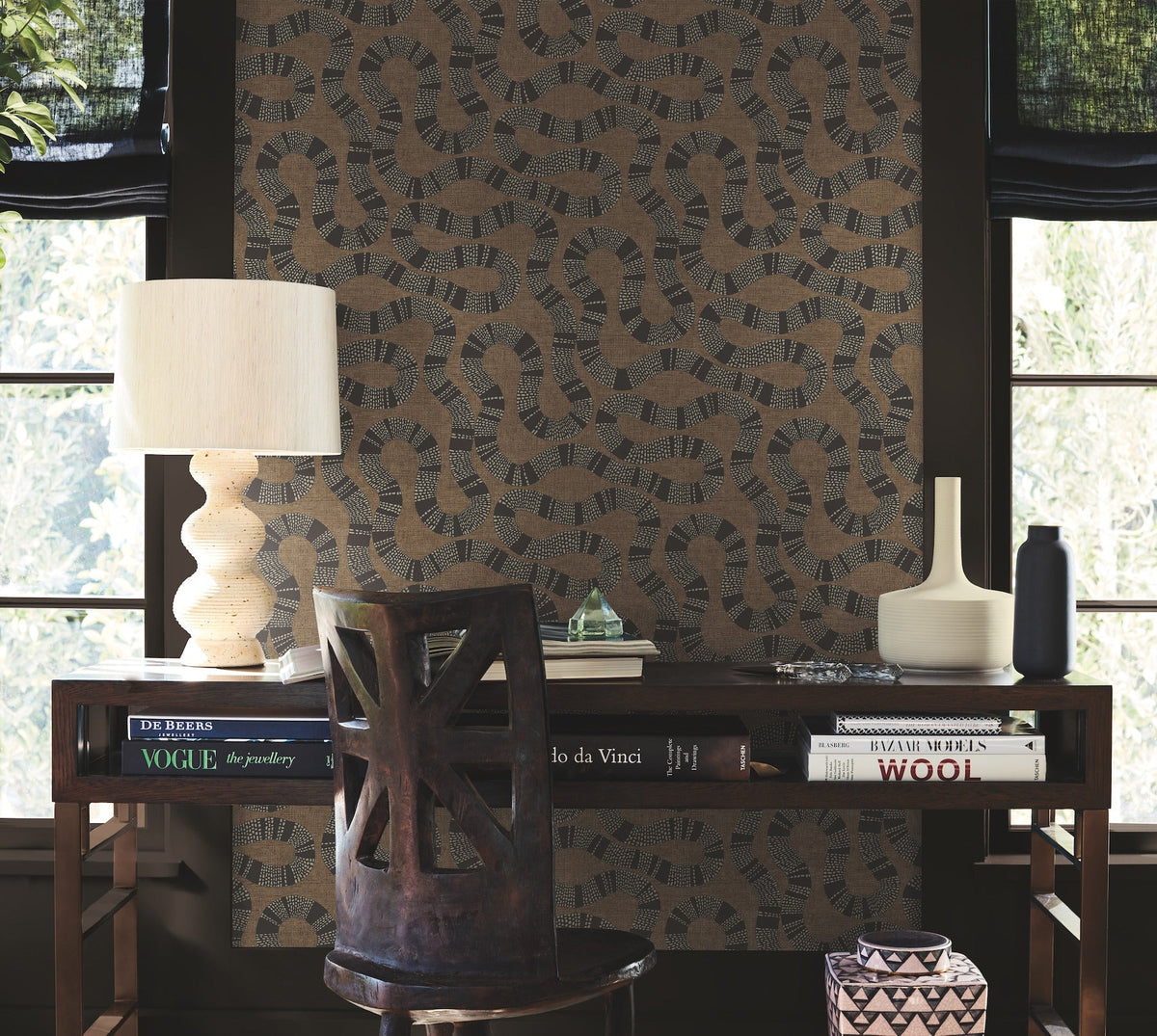 A wooden desk with books, a sculptural lamp, vase, and bottle sits in front of a window and a bold accent wall adorned with Ophidia Wallpaper by York Wallcoverings; a carved wooden chair is placed at the desk.