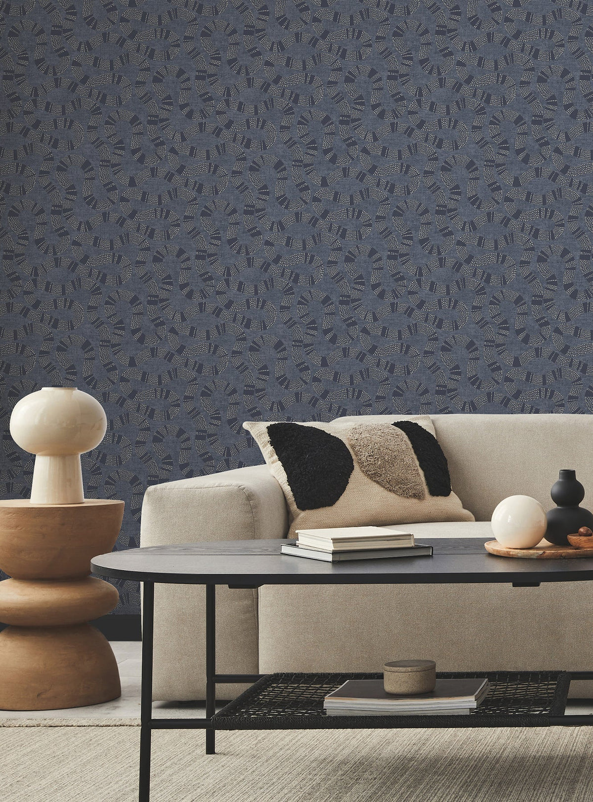 A modern living room with a light beige sofa, black and beige pillows, a round black coffee table, a sculptural beige side table with a lamp, and York Wallcoverings’ Ophidia Wallpaper featuring a blue pattern in the background.