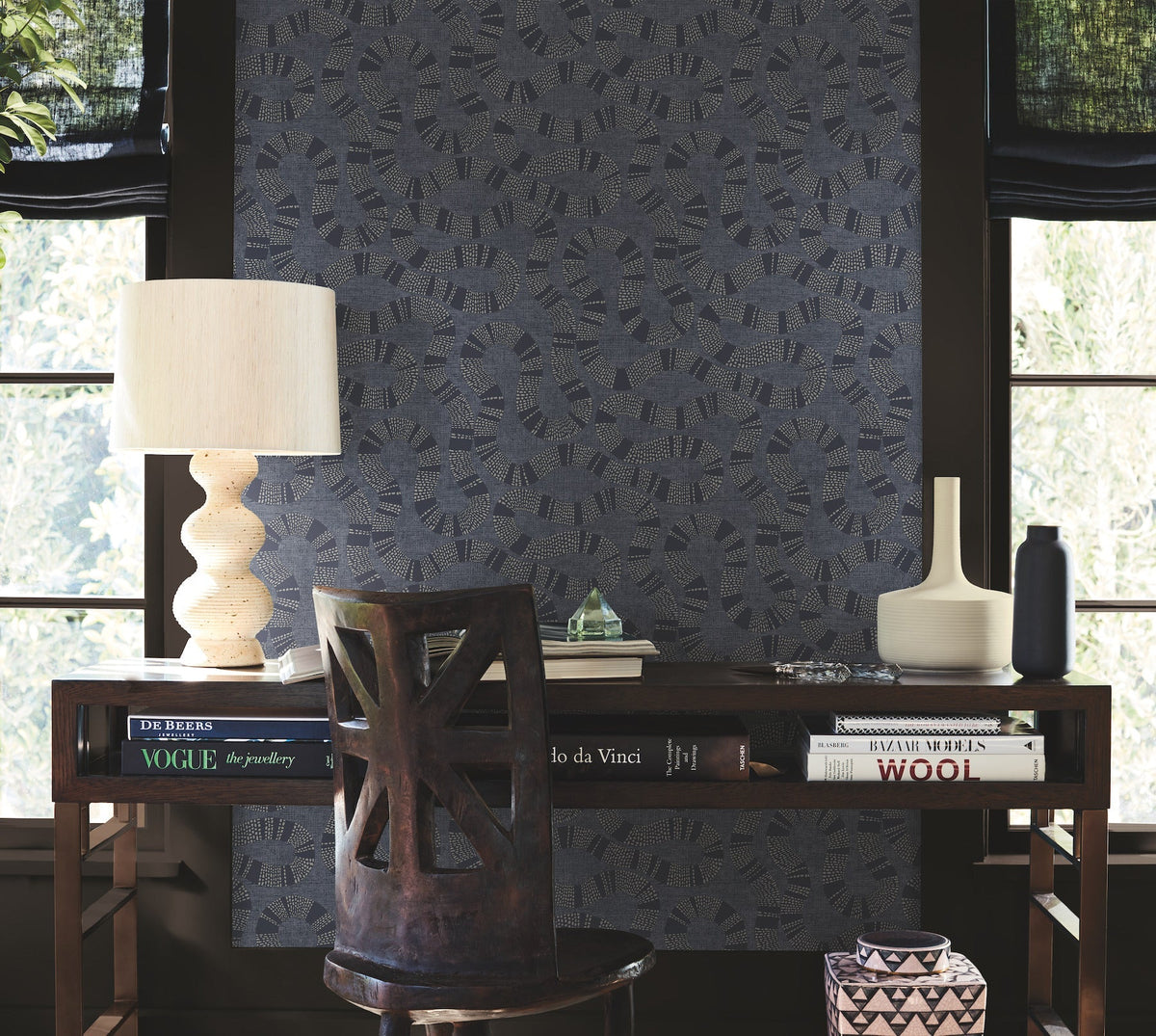 A desk with books, a sculptural lamp, a chair, and decorative vases sits in front of a window with black shades and York Wallcoverings’ Ophidia Wallpaper. Light filters in, giving the space a cozy, artistic vibe.