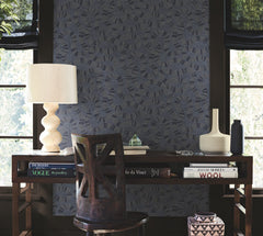 A desk with books, a sculptural lamp, a chair, and decorative vases sits in front of a window with black shades and York Wallcoverings’ Ophidia Wallpaper. Light filters in, giving the space a cozy, artistic vibe.