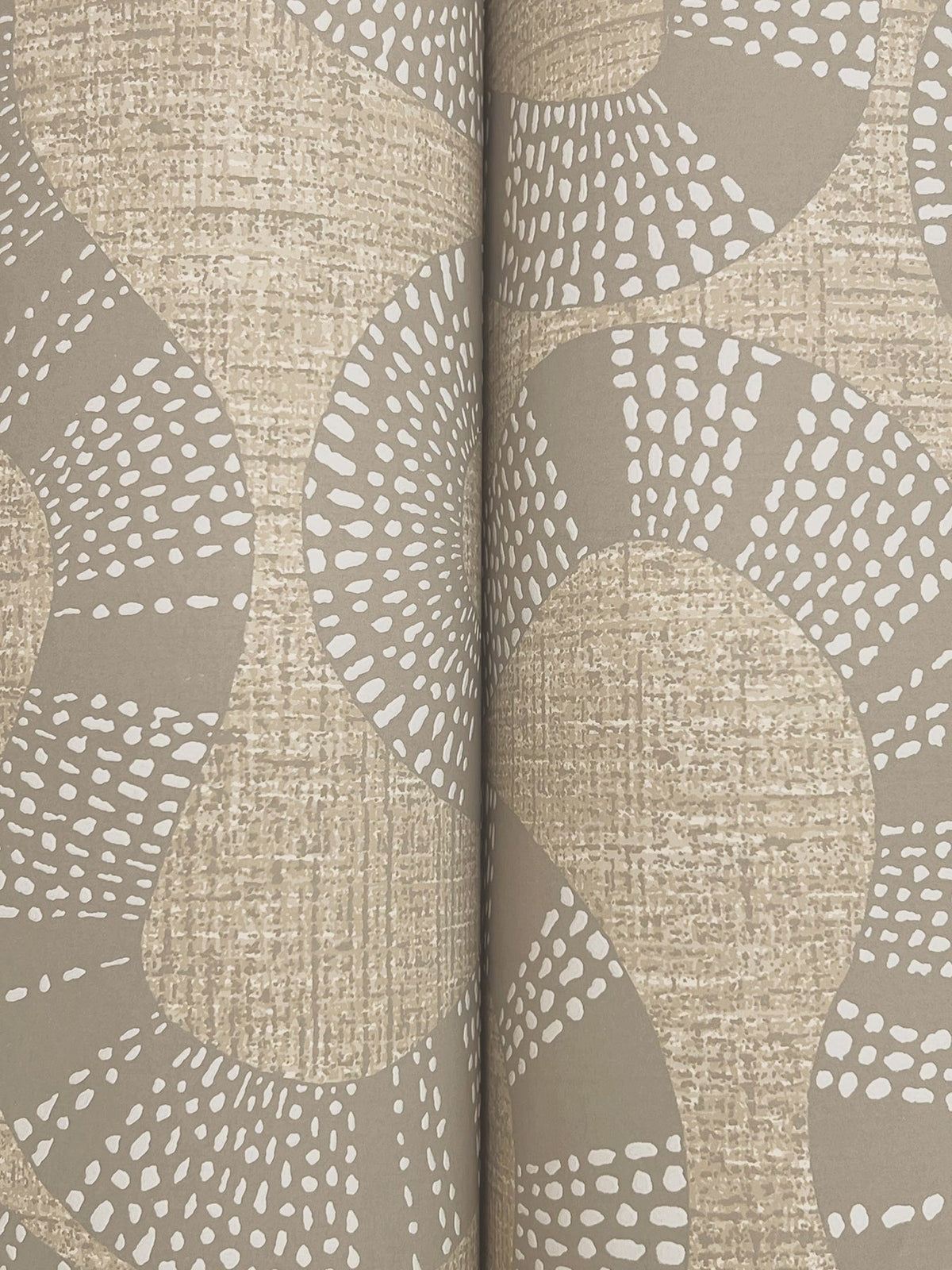 Close-up of York Wallcoverings’ Ophidia Wallpaper, featuring a tan and beige abstract pattern with curving lines and white dotted accents for added texture. Two rolls are placed side by side, forming a seam in the center.