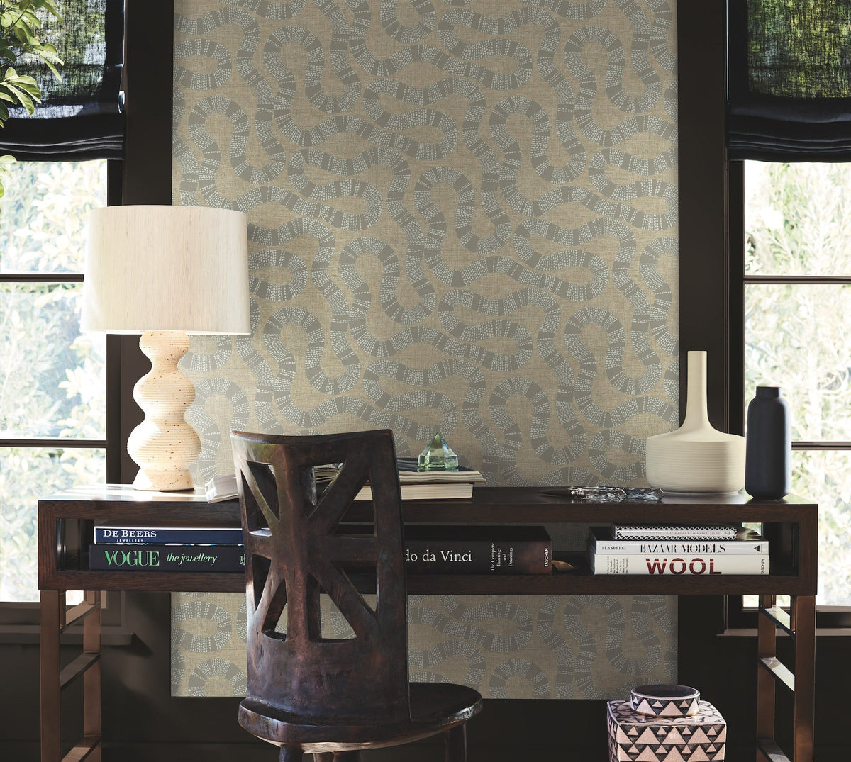 A desk with books, a lamp, vases, and a chair sits in front of two windows with black shades. The wall behind is adorned with Ophidia Wallpaper by York Wallcoverings, featuring a geometric pattern in beige and blue tones.