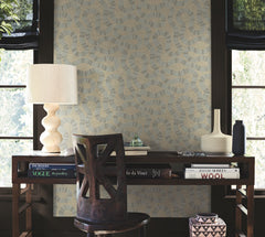 A desk with books, a lamp, vases, and a chair sits in front of two windows with black shades. The wall behind is adorned with Ophidia Wallpaper by York Wallcoverings, featuring a geometric pattern in beige and blue tones.