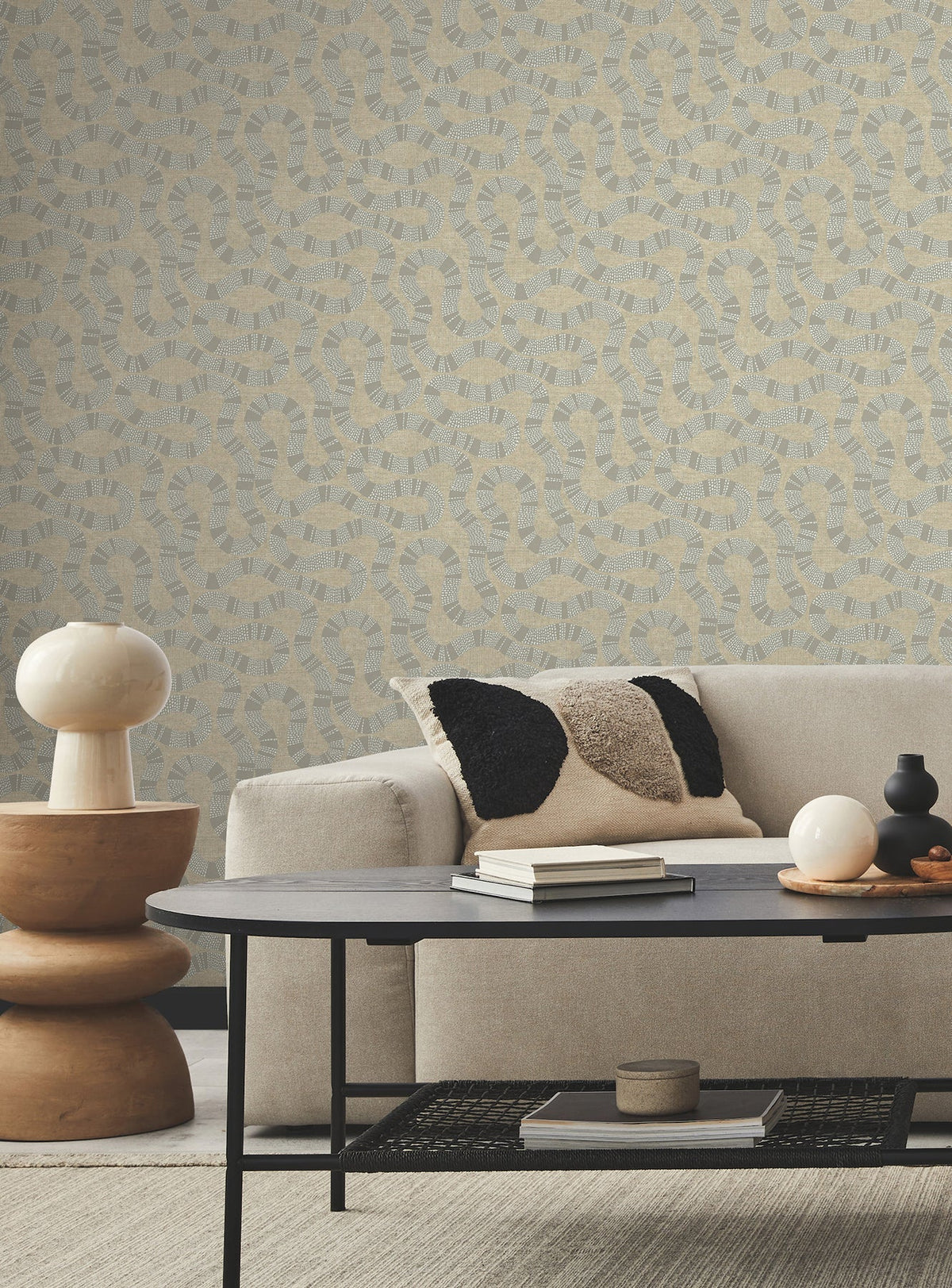 A modern living room featuring a light beige sofa, black and beige pillow, round wooden side table with a lamp, black coffee table, small decor items, and Ophidia Wallpaper by York Wallcoverings in neutral tones.