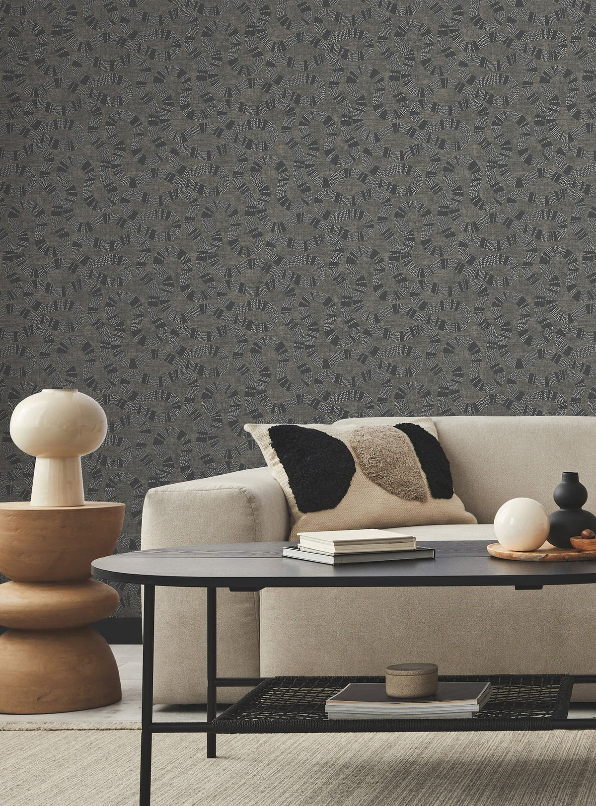A modern living room with a beige sofa, black-and-tan textured pillow, round table lamp on stacked wooden side tables, black coffee table, and Ophidia Wallpaper by York Wallcoverings in a patterned gray design.