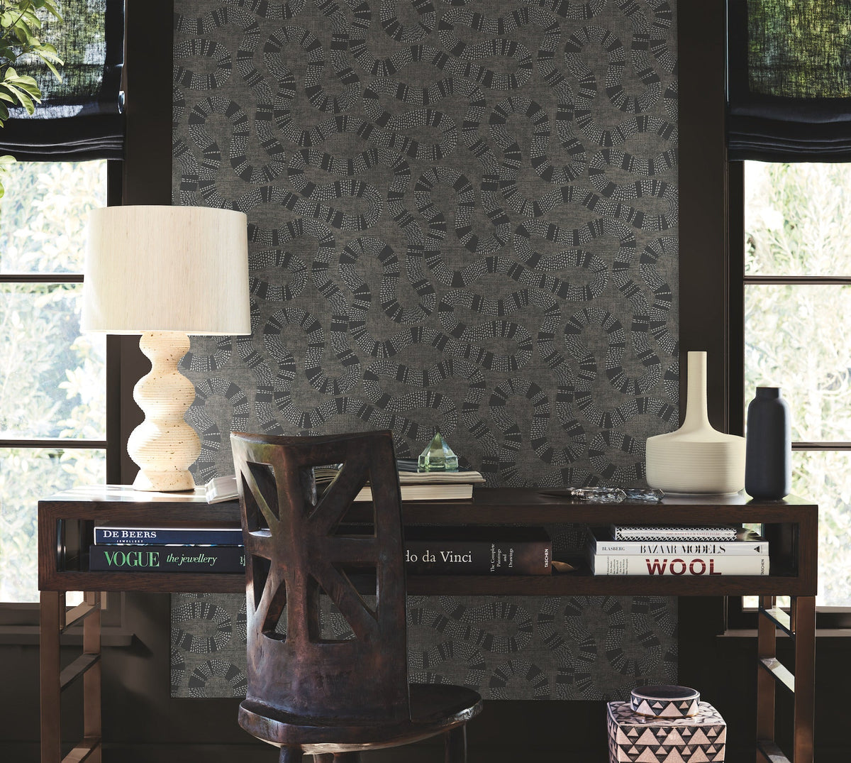 A wooden desk with books, a sculpted lamp, a vase, and a black bottle sits in front of two windows. A carved chair accompanies the desk, while York Wallcoverings’ Ophidia Wallpaper with its geometric pattern adorns the background wall.