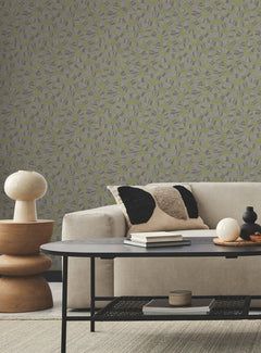 Modern living room with a beige sofa, black and beige pillows, round table, stacked wooden side table with a lamp, and textured abstract Ophidia Wallpaper in gray and green tones by York Wallcoverings. Cozy, minimalist decor.