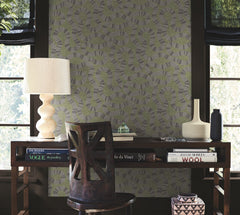 A wooden desk adorned with books, a beige lamp, vases, and paired with a wooden chair sits in front of York Wallcoverings’ Ophidia Wallpaper and two windows framed by black curtains, letting in natural light.