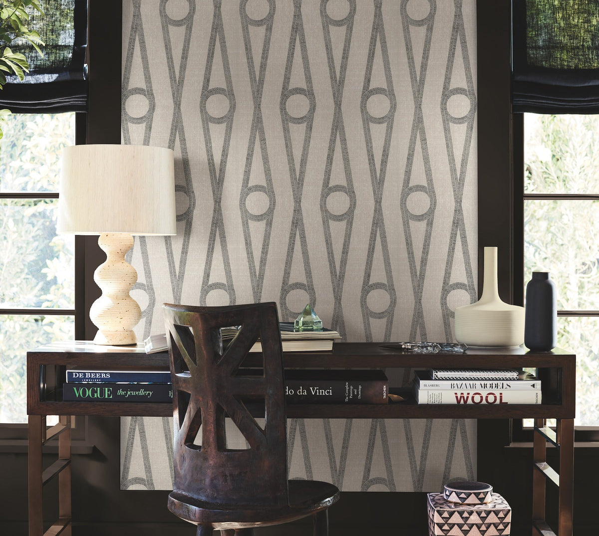Modern workspace featuring a dark wooden desk, decorative lamp, books, and vases. The chair showcases a distinctive cut-out back. Henlein Wallpaper by York Wallcoverings adds geometric interest to the background, complemented by large windows with black shades that allow light to filter in from the lush greenery outside.