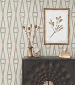 A dark wood cabinet with geometric patterns is topped with decor, including dried flowers, a crystal, and stacked books. A framed minimalist botanical print hangs on a wall covered in the Henlein Wallpaper by York Wallcoverings.