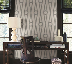 A modern desk with books, a sculptural lamp, and vases sits against a gray patterned wall covered in Henlein Wallpaper by York Wallcoverings between two windows. A wooden chair with a geometric back is placed at the desk.