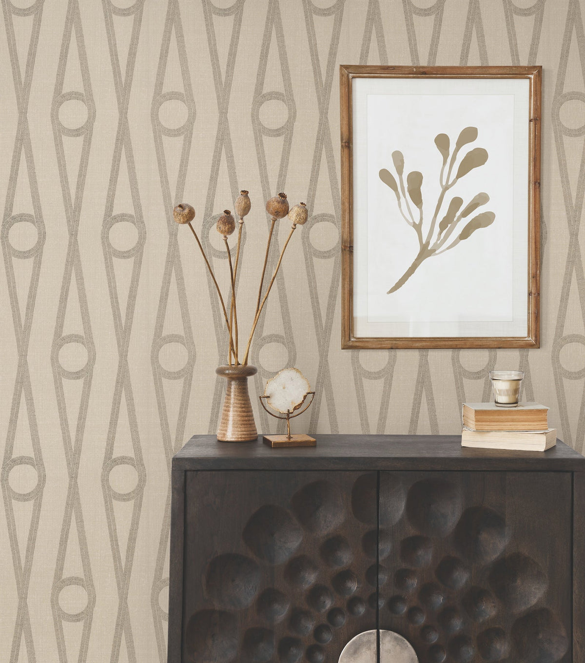 A dark wooden cabinet with a textured front sits against Henlein Wallpaper by York Wallcoverings, featuring a beige geometric pattern. On top are dried flowers in a vase, books, a cup, and a crystal. Above hangs framed minimalist botanical art.