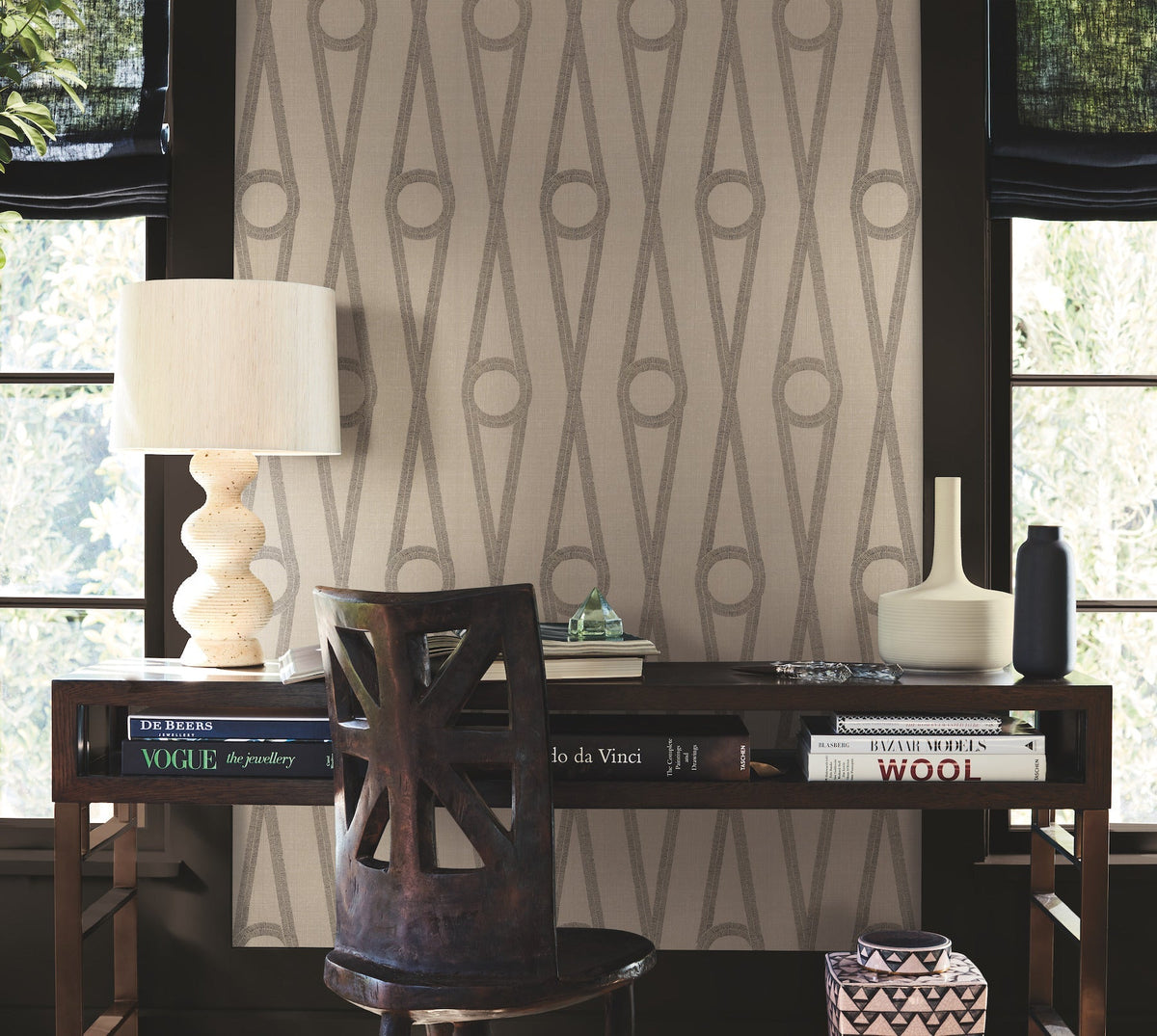A wooden desk topped with books, a table lamp, and vases is positioned in front of a window adorned with Henlein Wallpaper by York Wallcoverings. A dark wooden chair is tucked beneath the desk as light filters through the window.