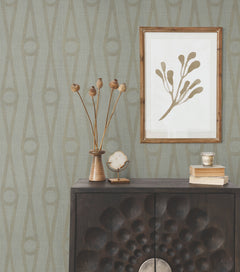 A modern cabinet with a textured front sits against Henlein Wallpaper by York Wallcoverings. On top are dried flowers, a crystal, books, and a metallic cup. Above, a framed minimalist leaf artwork hangs on the wall.