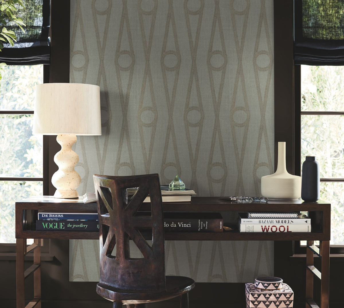A desk with books, a white lamp, two vases, and a wooden chair in front of two windows. The wall features Henlein Wallpaper by York Wallcoverings in a geometric pattern with neutral tones.