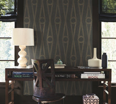 A dark wooden desk with books, a sculpted lamp, and vases sits in front of a window and a wall adorned with Henlein Wallpaper by York Wallcoverings. A wooden chair is pushed under the desk as sunlight filters through the windows and blinds.