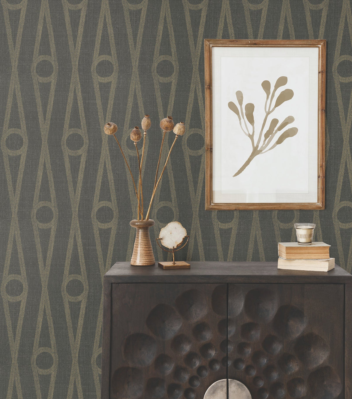 A dark wood cabinet with a textured front stands against York Wallcoverings’ Henlein Wallpaper. On top are dried flowers, books, and a crystal, while a framed botanical print hangs above.