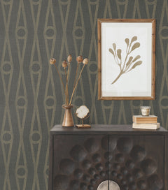A dark wood cabinet with a carved pattern holds vases, books, and decor. Above it, a framed abstract plant print hangs on a wall covered in Henlein Wallpaper by York Wallcoverings, featuring retro geometric patterns in muted tones.