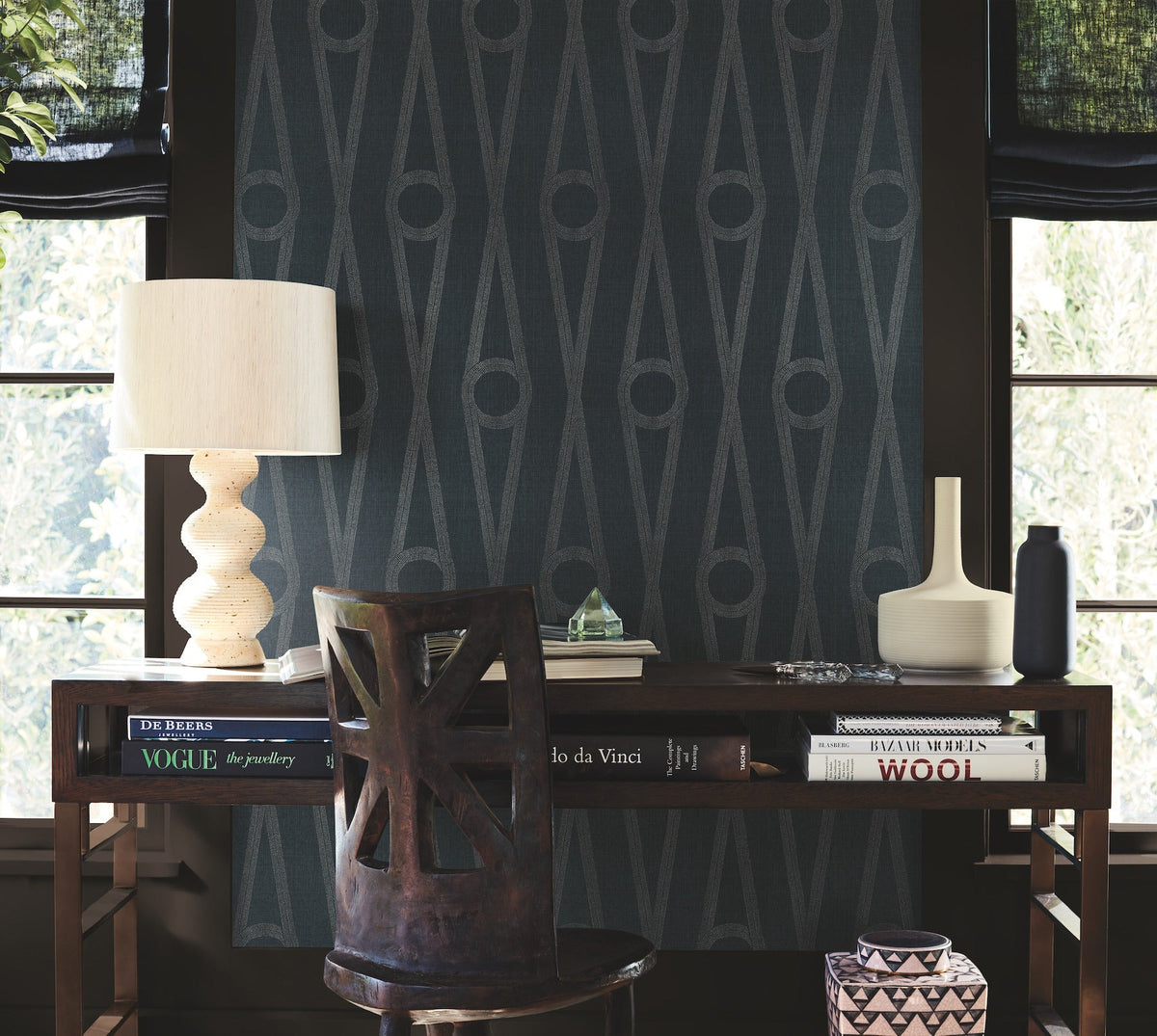 A wooden desk topped with books, a lamp, and vases sits in front of York Wallcoverings’ Henlein Wallpaper, featuring modern geometric patterns. A chair with a cut-out back is pushed in, while large windows frame the cozy workspace.