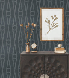A dark wood cabinet with textured doors is styled with dried flowers in a vase, books, a crystal, and a cup. Above, a framed minimalist plant print hangs on a wall adorned with Henlein Wallpaper by York Wallcoverings in rich dark blue.