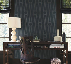 In front of the window, a desk styled with books, decorative vases, a sculpted lamp, and a cutout-back chair is set against Henlein Wallpaper by York Wallcoverings, with natural light streaming in.