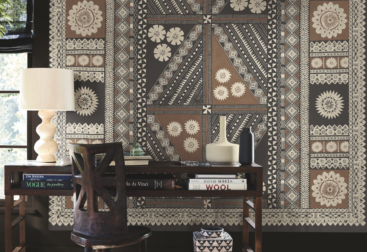 A desk with books, a lamp, and vases sits in front of a wall adorned with the Tapa Mural Wall Mural, featuring an intricate geometric and floral pattern in beige, brown, and black tones.