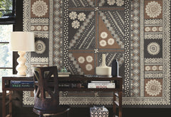 A desk with books, a lamp, and vases sits in front of a wall adorned with the Tapa Mural Wall Mural, featuring an intricate geometric and floral pattern in beige, brown, and black tones.