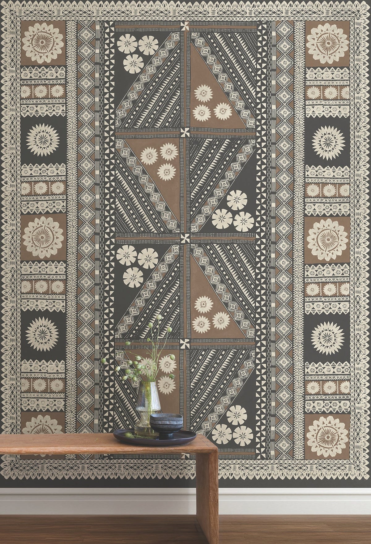 A Tapa Mural Wall Mural featuring geometric shapes and floral motifs in black, brown, and cream tones is displayed with a wooden bench in front, adorned with vases and books.