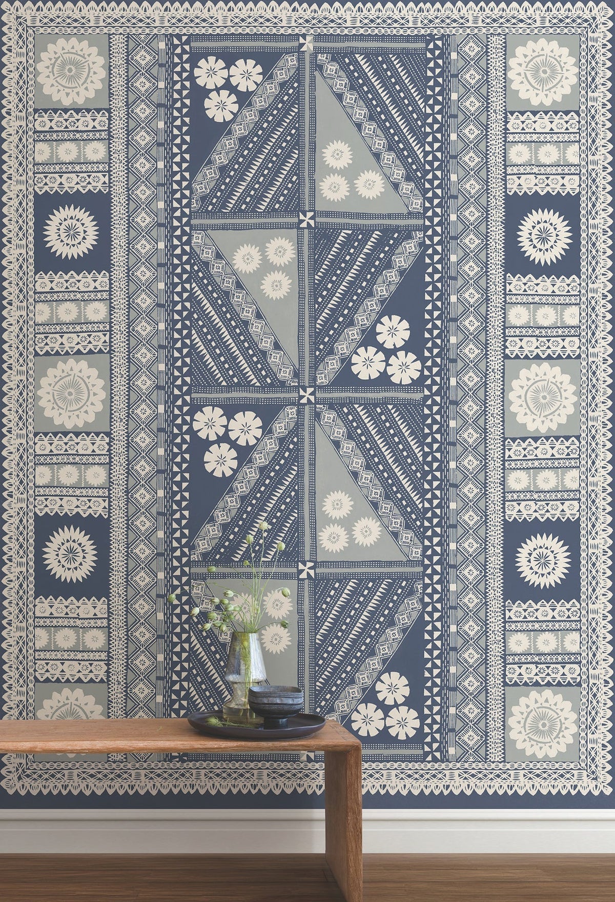 The Tapa Mural Wall Mural featuring blue and white floral and geometric patterns is displayed behind a wooden bench that holds a glass vase with flowers and a stack of books.