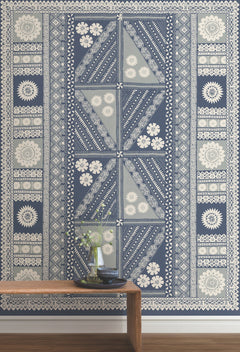 The Tapa Mural Wall Mural featuring blue and white floral and geometric patterns is displayed behind a wooden bench that holds a glass vase with flowers and a stack of books.
