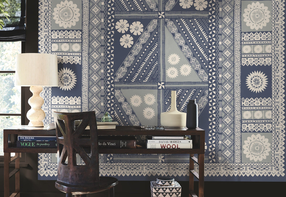 A wooden desk with books, ceramics, and a lamp sits in front of a wall adorned with the Tapa Mural Wall Mural, featuring blue and white geometric and floral patterns. Sunlight filters in from a window on the left.