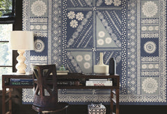 A wooden desk with books, ceramics, and a lamp sits in front of a wall adorned with the Tapa Mural Wall Mural, featuring blue and white geometric and floral patterns. Sunlight filters in from a window on the left.